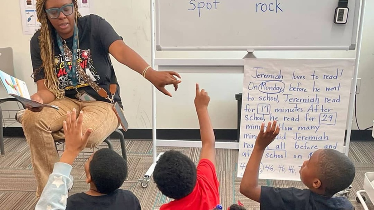 Teacher reading aloud to a group of students who raise their hands in a classroom during a read-aloud lesson with a reading poster nearby.