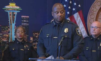 Black police officer speaks at a podium during a press conference, flanked by two officers, with the Seattle skyline behind.