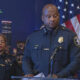 Black police officer speaks at a podium during a press conference, flanked by two officers, with the Seattle skyline behind.