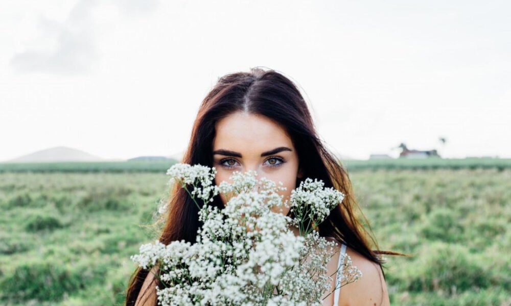 Woman in a field holding white baby's breath flowers in front of her face, revealing her blue eyes.