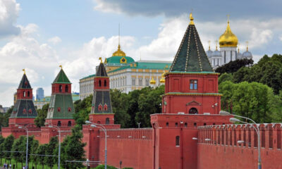 Red brick Kremlin wall with green-roofed towers stretching along a street, green trees behind and gold-domed churches in the background. This image shows the Moscow Kremlin complex.