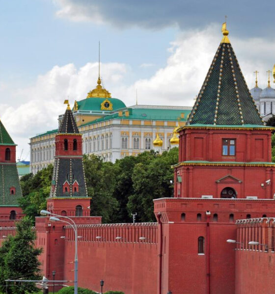 Red brick Kremlin wall with green-roofed towers stretching along a street, green trees behind and gold-domed churches in the background. This image shows the Moscow Kremlin complex.