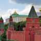 Red brick Kremlin wall with green-roofed towers stretching along a street, green trees behind and gold-domed churches in the background. This image shows the Moscow Kremlin complex.