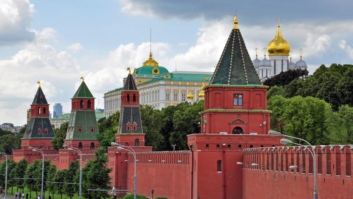 Red brick Kremlin wall with green-roofed towers stretching along a street, green trees behind and gold-domed churches in the background. This image shows the Moscow Kremlin complex.