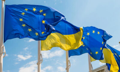 EU and Ukraine flags waving in the wind against a bright blue sky; the EU flag with yellow stars on blue and the Ukrainian flag with blue over yellow stripes partially overlapping.