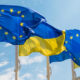 EU and Ukraine flags waving in the wind against a bright blue sky; the EU flag with yellow stars on blue and the Ukrainian flag with blue over yellow stripes partially overlapping.