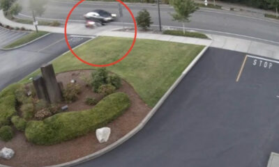 Blurred motorcycle or scooter moving across a sidewalk near a landscaped median by a parking area and road intersection.