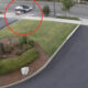 Blurred motorcycle or scooter moving across a sidewalk near a landscaped median by a parking area and road intersection.