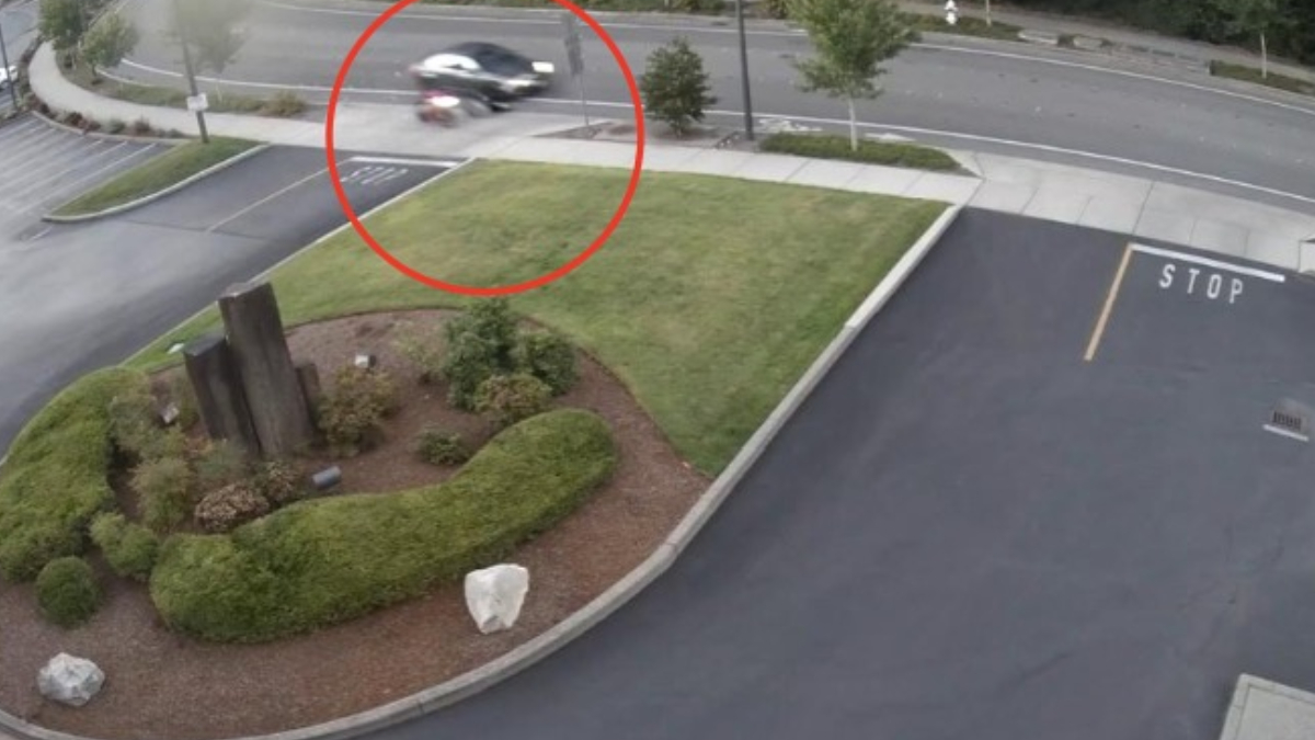 Blurred motorcycle or scooter moving across a sidewalk near a landscaped median by a parking area and road intersection.