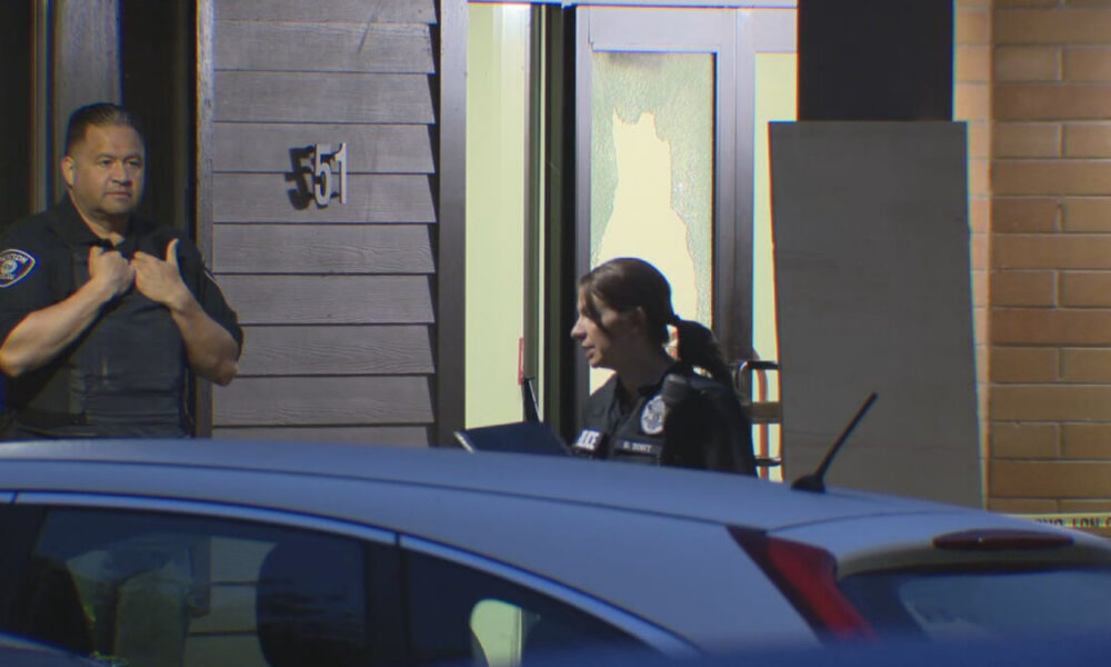 Two police officers stand outside a building, one man adjusting his vest while a woman officer occupies the doorway, with a damaged window behind them and a blue car in the foreground.