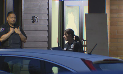 Two police officers stand outside a building, one man adjusting his vest while a woman officer occupies the doorway, with a damaged window behind them and a blue car in the foreground.