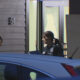 Two police officers stand outside a building, one man adjusting his vest while a woman officer occupies the doorway, with a damaged window behind them and a blue car in the foreground.