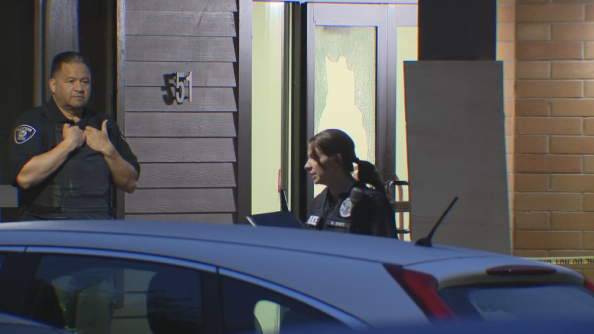 Two police officers stand outside a building, one man adjusting his vest while a woman officer occupies the doorway, with a damaged window behind them and a blue car in the foreground.