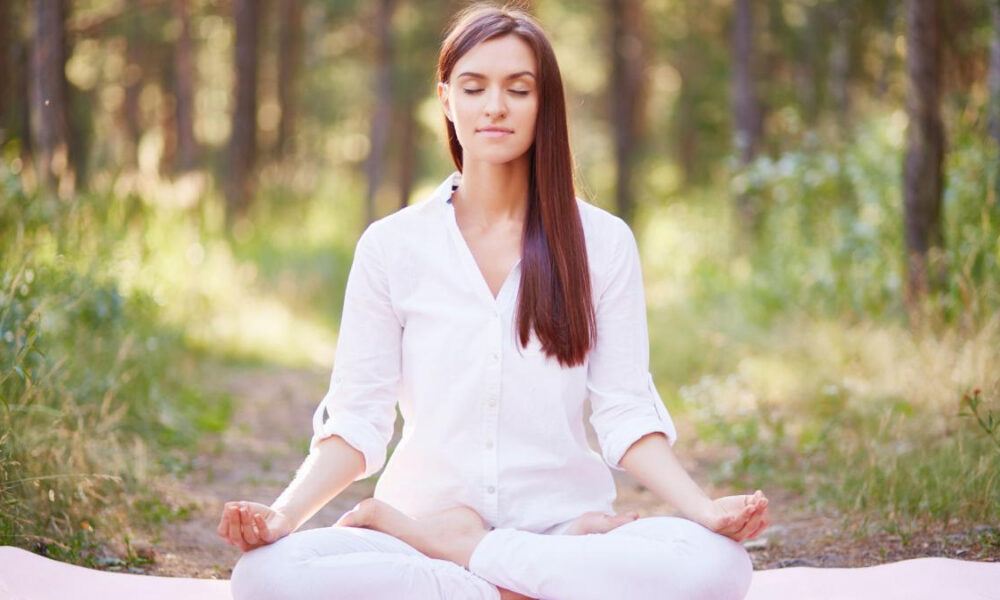 Woman meditating in a forest, seated on a mat with legs crossed and hands resting on knees, eyes closed in calm posture.