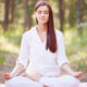 Woman meditating in a forest, seated on a mat with legs crossed and hands resting on knees, eyes closed in calm posture.
