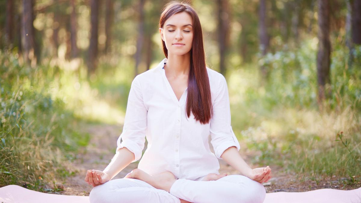 Woman meditating in a forest, seated on a mat with legs crossed and hands resting on knees, eyes closed in calm posture.