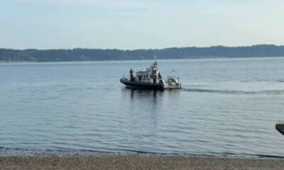 Small tugboat towing a small boat across a calm bay, with a tree-lined shoreline in the distance and a pebbly beach in the foreground.