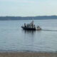 Small tugboat towing a small boat across a calm bay, with a tree-lined shoreline in the distance and a pebbly beach in the foreground.