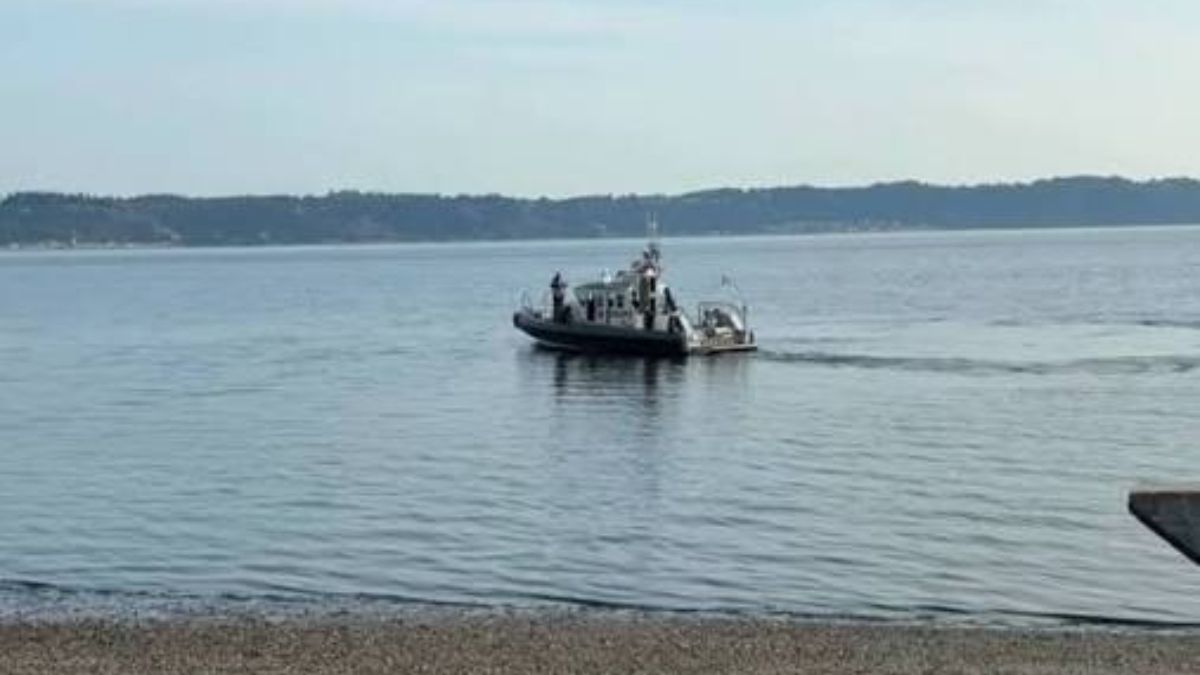 Small tugboat towing a small boat across a calm bay, with a tree-lined shoreline in the distance and a pebbly beach in the foreground.