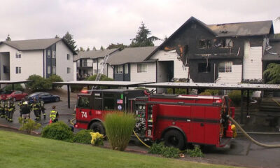 Firefighters and a red fire engine battle a two-story, burned apartment building with charred upper floor and smoke lingering. A ladder leans against the structure.