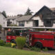 Firefighters and a red fire engine battle a two-story, burned apartment building with charred upper floor and smoke lingering. A ladder leans against the structure.