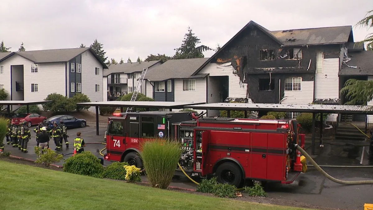 Firefighters and a red fire engine battle a two-story, burned apartment building with charred upper floor and smoke lingering. A ladder leans against the structure.