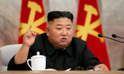 A man in a dark suit at a conference table, raising one finger while speaking, with red flags behind him and a white teacup on the table.