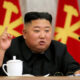 A man in a dark suit at a conference table, raising one finger while speaking, with red flags behind him and a white teacup on the table.