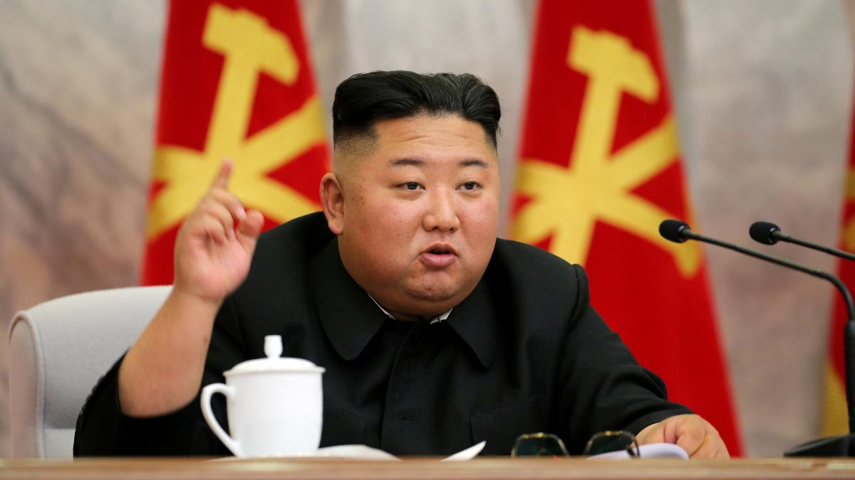 A man in a dark suit at a conference table, raising one finger while speaking, with red flags behind him and a white teacup on the table.