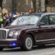 Maroon luxury sedan driving past a crowd of spectators on a city street; circular inset shows a man in a blue suit portrait.
