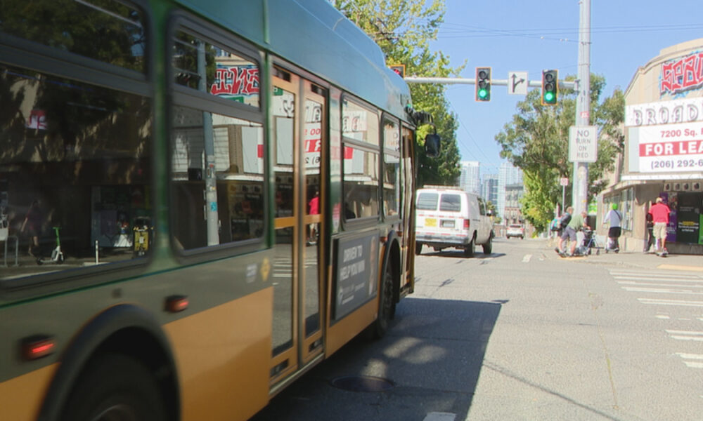 City street with a green-yellow bus at a crosswalk, cars ahead, pedestrians near storefronts including a 'For Lease' sign.