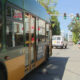 City street with a green-yellow bus at a crosswalk, cars ahead, pedestrians near storefronts including a 'For Lease' sign.