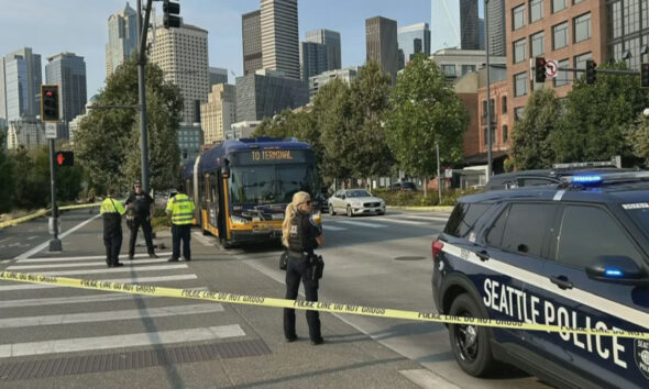 Seattle street scene with police tape and officers guarding a bus near downtown skyline.