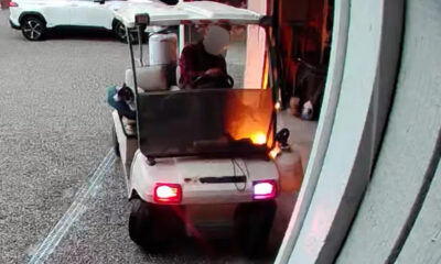 White golf cart on a parking lot, engine area aflame with visible orange flames as it moves away.