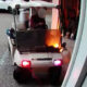White golf cart on a parking lot, engine area aflame with visible orange flames as it moves away.