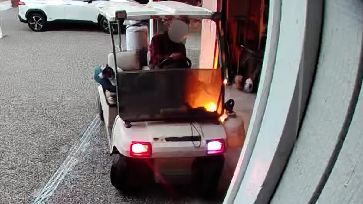 White golf cart on a parking lot, engine area aflame with visible orange flames as it moves away.