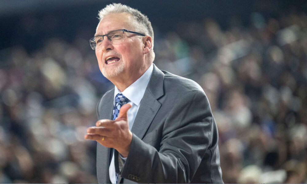 Man in a gray suit and blue tie speaks passionately to a large, blurred audience behind him.