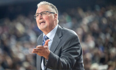 Man in a gray suit and blue tie speaks passionately to a large, blurred audience behind him.