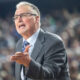 Man in a gray suit and blue tie speaks passionately to a large, blurred audience behind him.