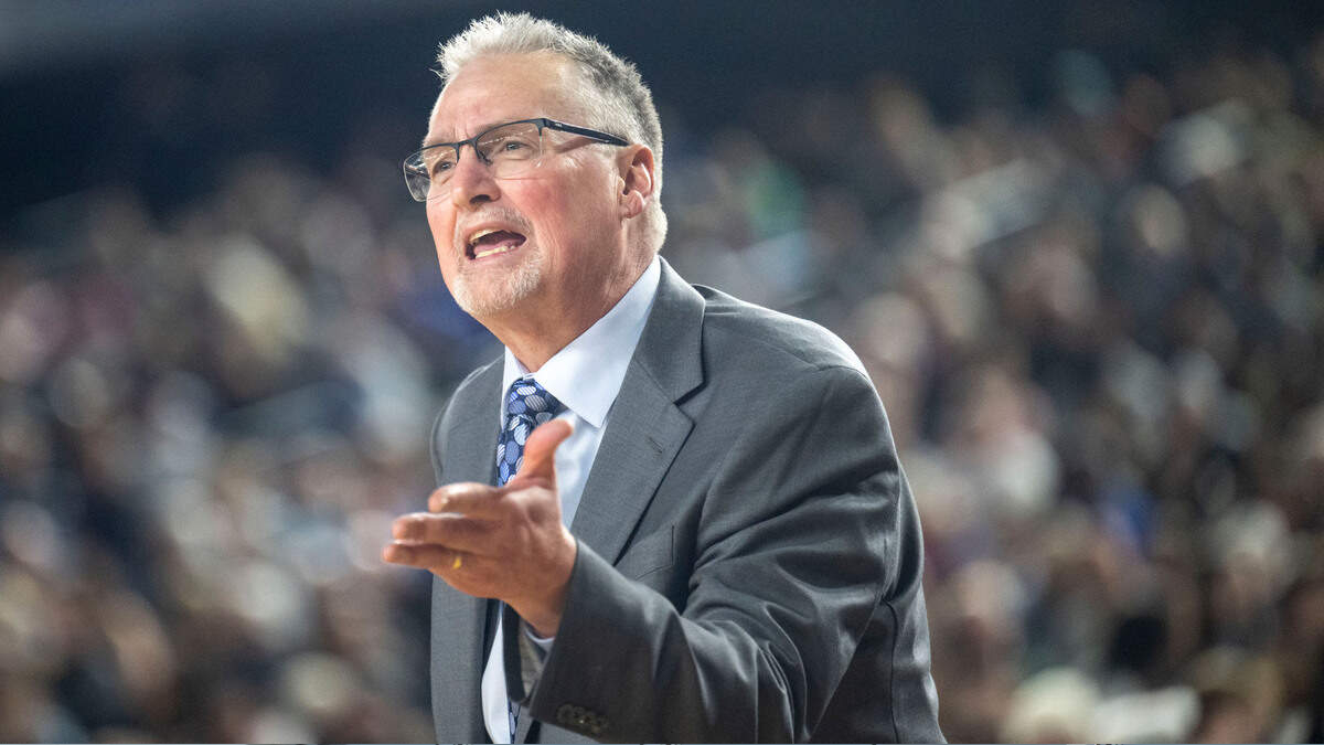 Man in a gray suit and blue tie speaks passionately to a large, blurred audience behind him.