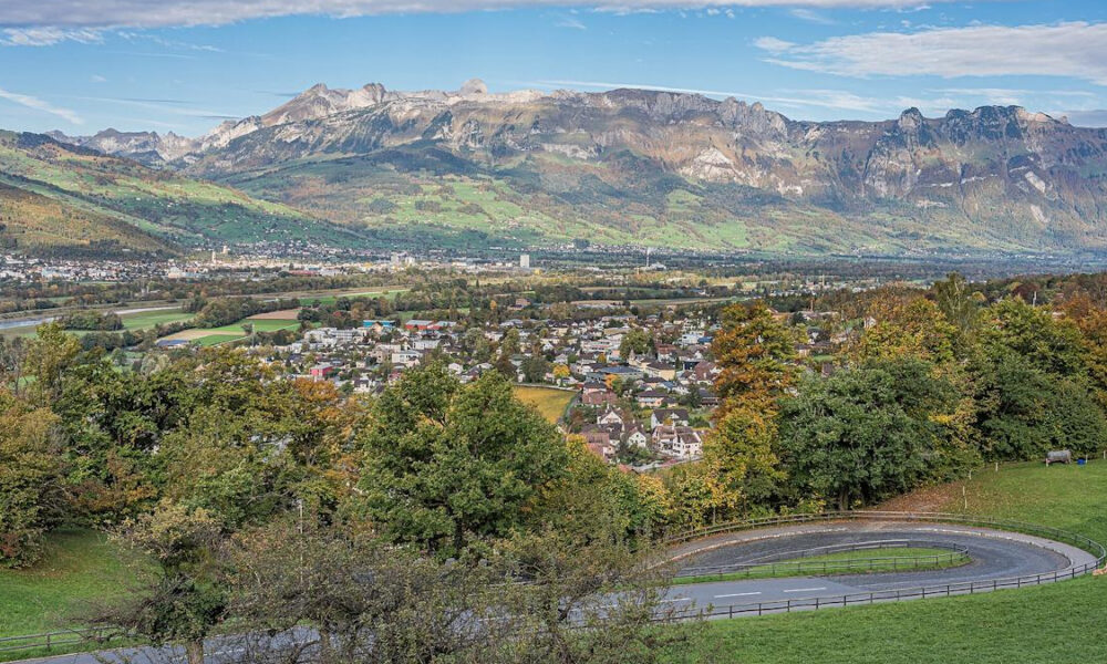 Panoramic valley town with a curved road in the foreground, forested hills, and distant rugged mountains under a blue sky.