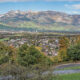 Panoramic valley town with a curved road in the foreground, forested hills, and distant rugged mountains under a blue sky.