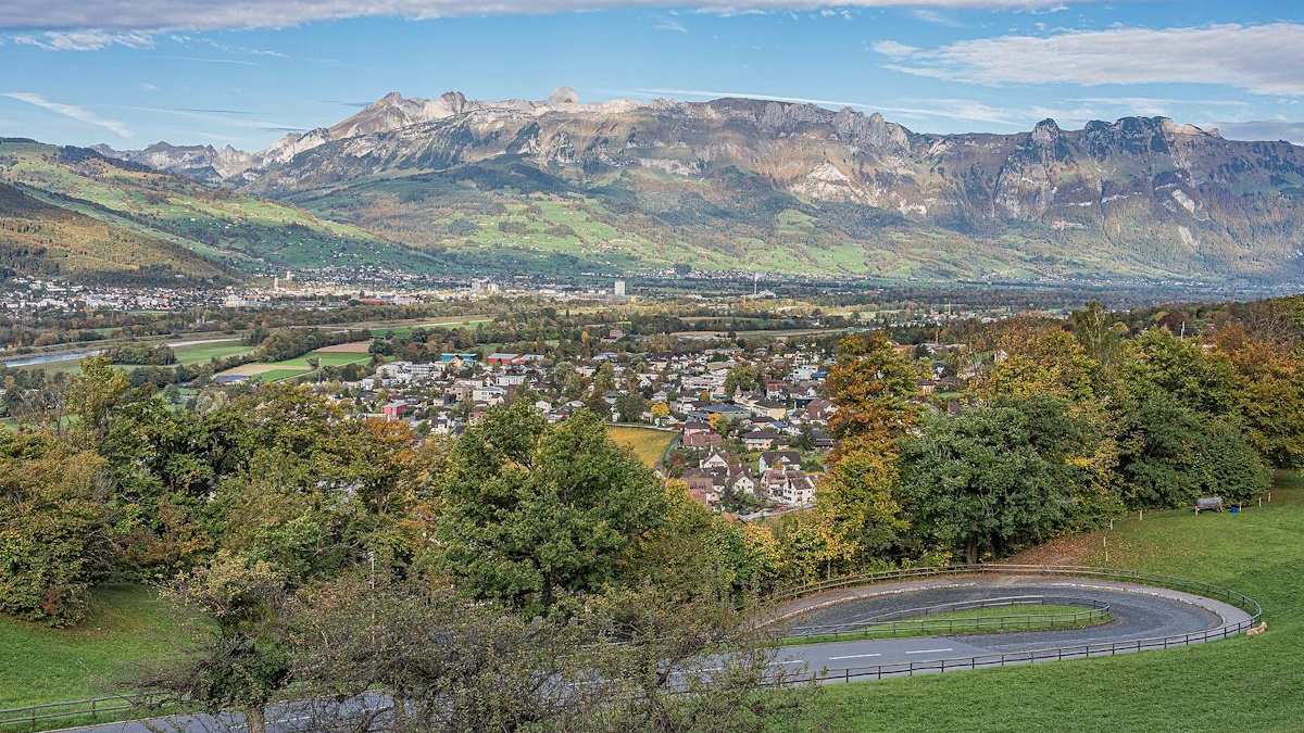 Panoramic valley town with a curved road in the foreground, forested hills, and distant rugged mountains under a blue sky.