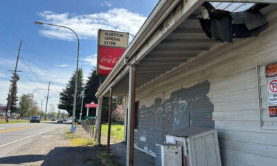 Dilapidated wooden storefront with peeling paint and graffiti, Alderton General Store sign and a red Coca‑Cola sign beside the road nearby.