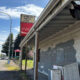 Dilapidated wooden storefront with peeling paint and graffiti, Alderton General Store sign and a red Coca‑Cola sign beside the road nearby.