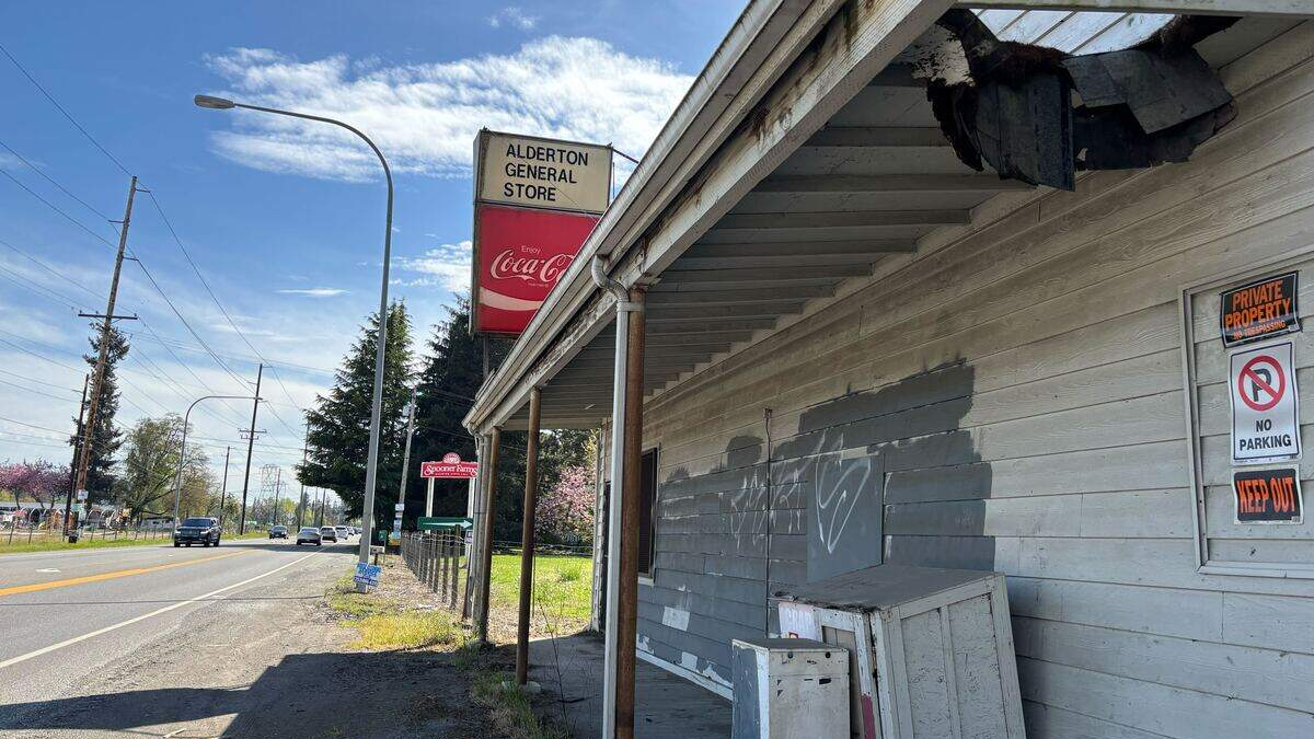 Dilapidated wooden storefront with peeling paint and graffiti, Alderton General Store sign and a red Coca‑Cola sign beside the road nearby.