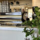 Shiny stainless espresso machine on a cafe counter with a stack of disposable cups and syrups behind it, and a leafy potted plant in the foreground.