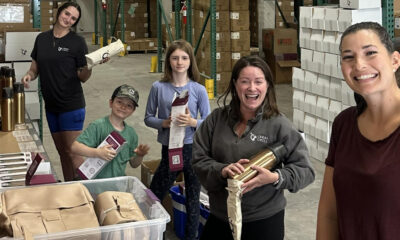 Smiling volunteers in a warehouse packaging supplies for donation or charity drive, with boxes and gear in the background.