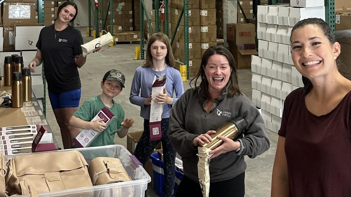 Smiling volunteers in a warehouse packaging supplies for donation or charity drive, with boxes and gear in the background.