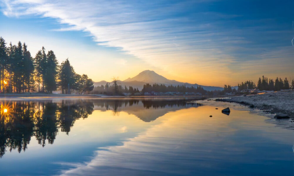 Sunrise over a calm lake with a snow-capped mountain and pine forest reflected in the water.